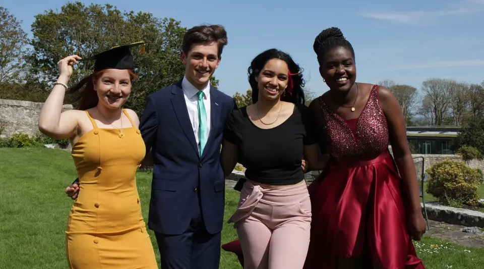Quattro persone sono insieme all'aperto nel campus del UWC Atlantic College con alberi e un cielo limpido sullo sfondo.