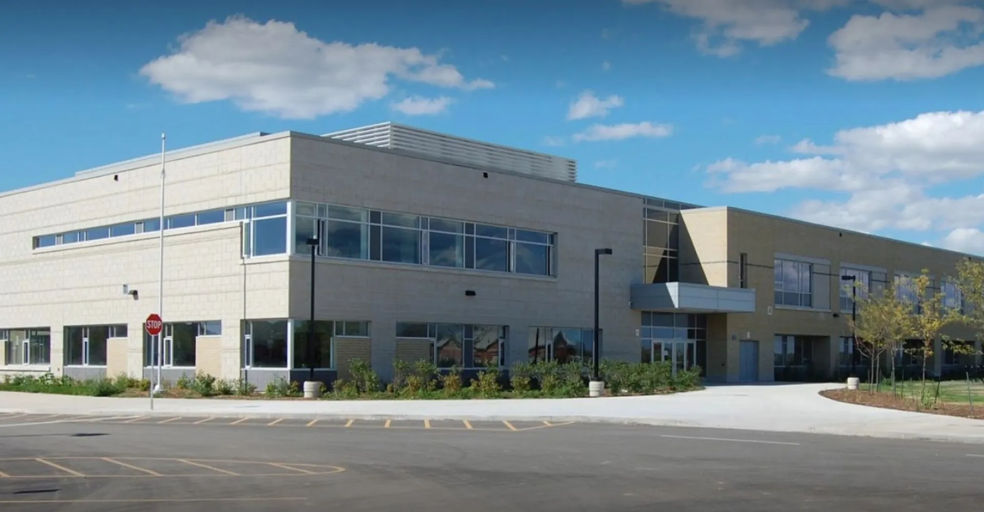 L'edificio moderno a più piani della Craig Kielburger Secondary School, con grandi finestre, si trova su un parcheggio pavimentato sotto un cielo blu.