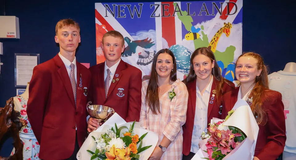 Un gruppo di studenti e studentesse dell'Inglewood High School in uniformi rosse con fiori e un trofeo davanti a una grande bandiera neozelandese.