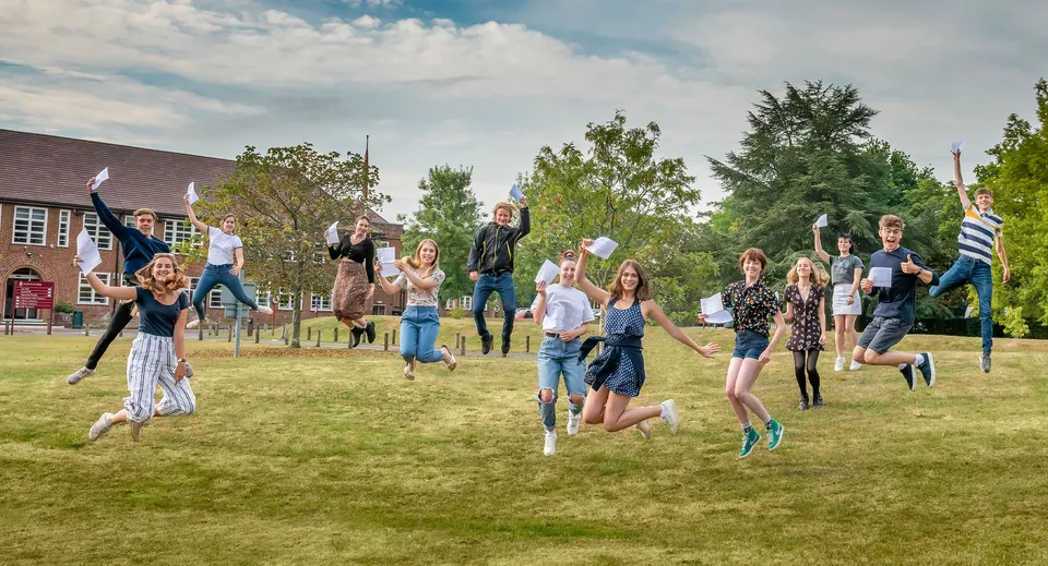 Un gruppo di studenti del Brockenhurst College salta e festeggia su un prato davanti ad alberi ed edifici.