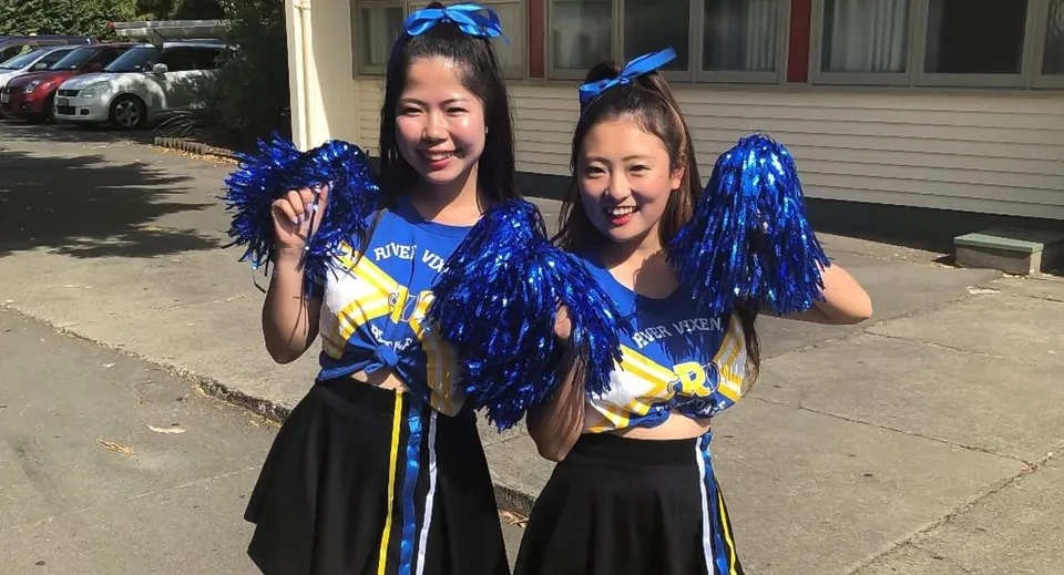 Due studentesse in divisa da cheerleader sono in piedi con i pompon davanti a un edificio del campus del Waimea College.