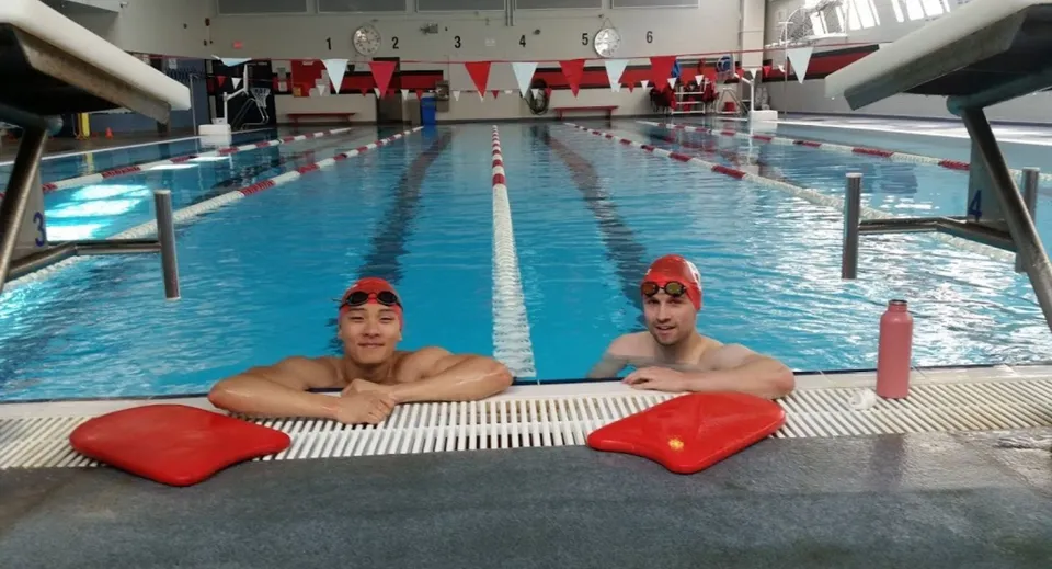 Due studenti con cuffie da nuoto rosse sono seduti su elementi galleggianti rossi nella piscina della St. George’s School.