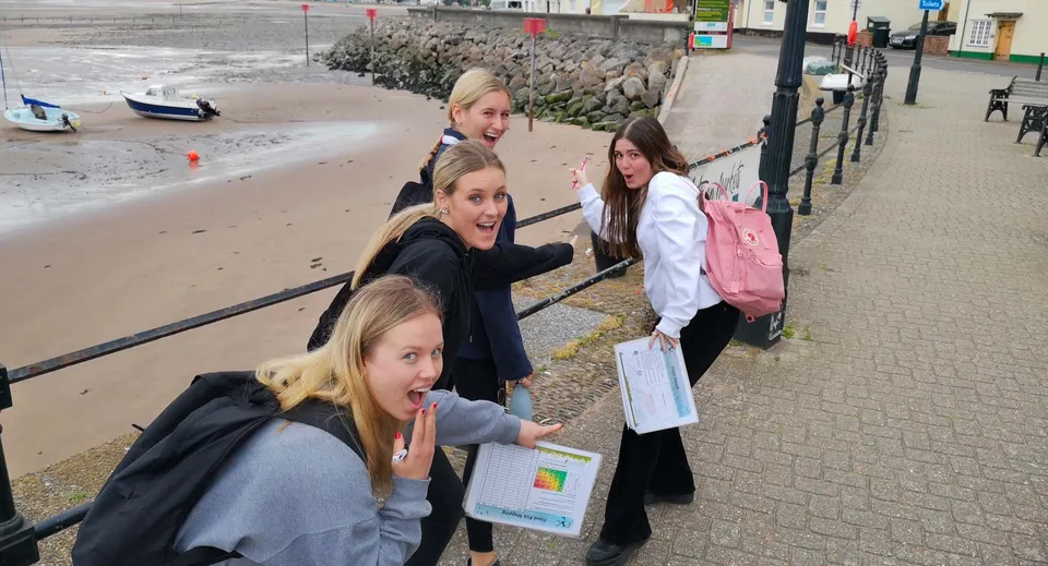 Un gruppo di studentesse della St. George's Ascot sta insieme su un marciapiede vicino a uno specchio d'acqua con una costa rocciosa ed edifici sullo sfondo.