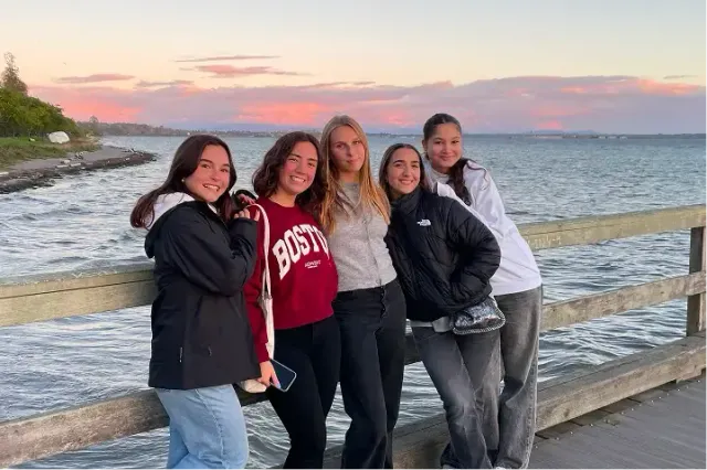 Cinque studentesse sorridono su un molo di legno al tramonto in riva al mare