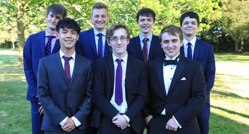 Un grupo de jóvenes en ropa formal está de pie en el campus de Padworth College al aire libre frente a árboles.