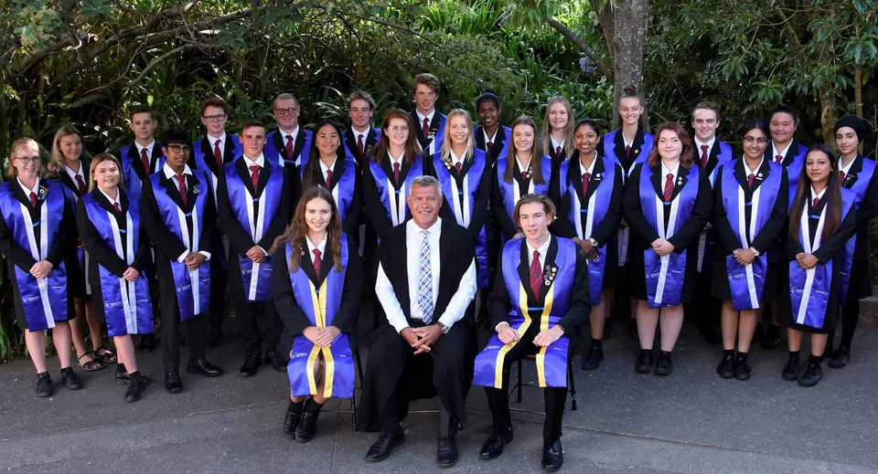 Un gruppo di diplomati dell'Howick College si trova insieme in abiti accademici all'aperto nel verde.