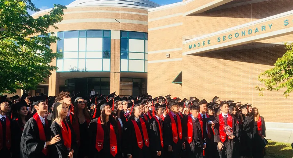 Un grande gruppo di diplomati e diplomate in toga rossa e nera davanti all'edificio in mattoni della Magee Secondary School.