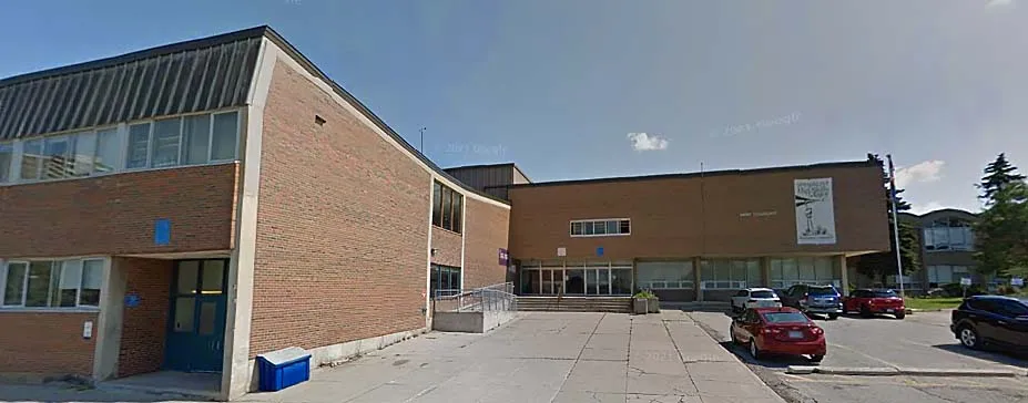 El moderno edificio de ladrillo con grandes ventanas del Emery Collegiate Institute está rodeado por una zona pavimentada con coches aparcados.