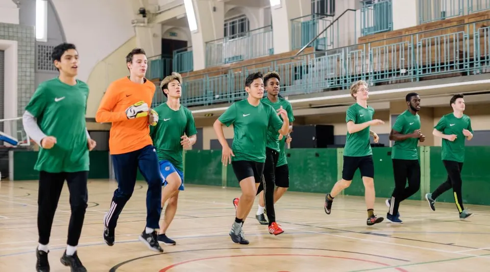 Un gruppo di studenti del David Game College Liverpool corre insieme nella palestra con campo da basket e tribune.