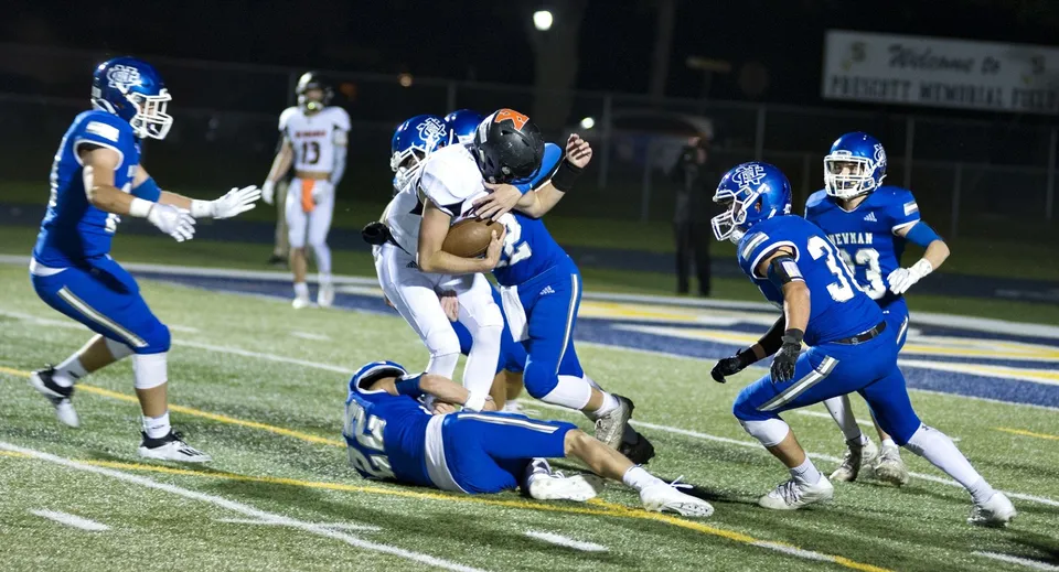 Giocatori della Newman Central Catholic High School visibili in un tackle sul campo durante una partita di football.