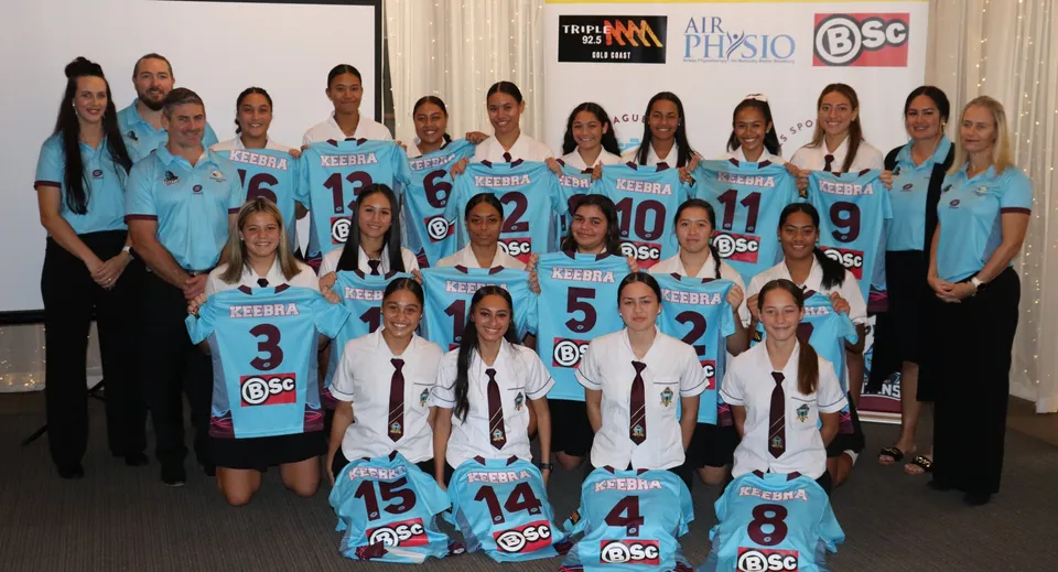Un gruppo di studentesse della Keebra Park State High School posa in divisa sportiva davanti a un pannello pubblicitario.