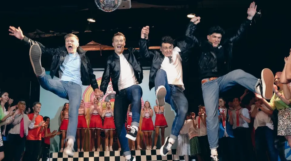 Un gruppo di studenti della Kinsale Community School si trova su un palco a scacchiera con uno sfondo decorato.