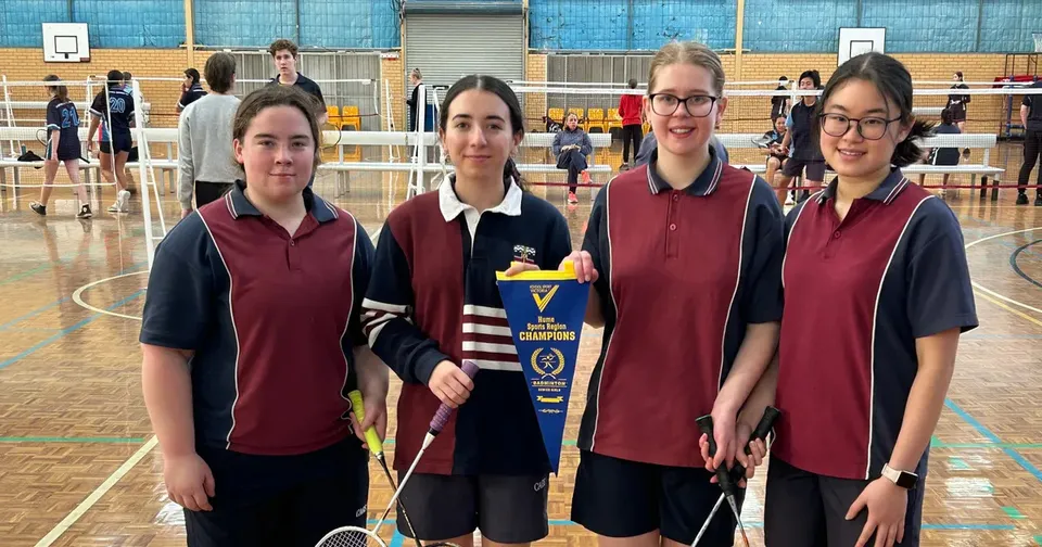 Quattro studentesse della Cobram Anglican Grammar School posano con racchette da badminton e un gagliardetto in una palestra.