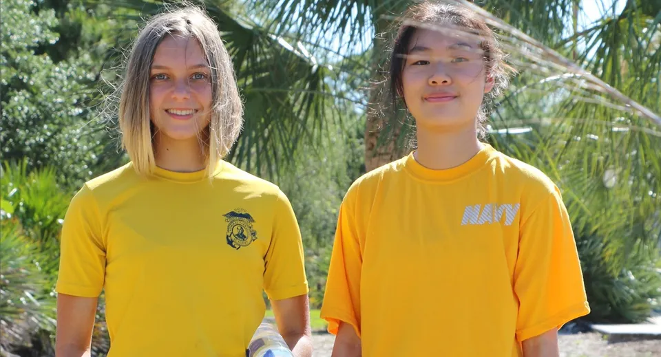 Due studenti della Admiral Farragut Academy indossano camicie giallo brillante e si trovano davanti a palme in un ambiente tropicale.