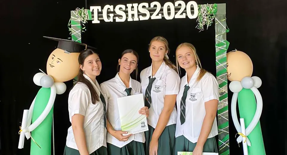 Un gruppo di quattro studentesse sta in piedi davanti a uno sfondo con la scritta TGSHS2020 nel campus della The Gap State High School.
