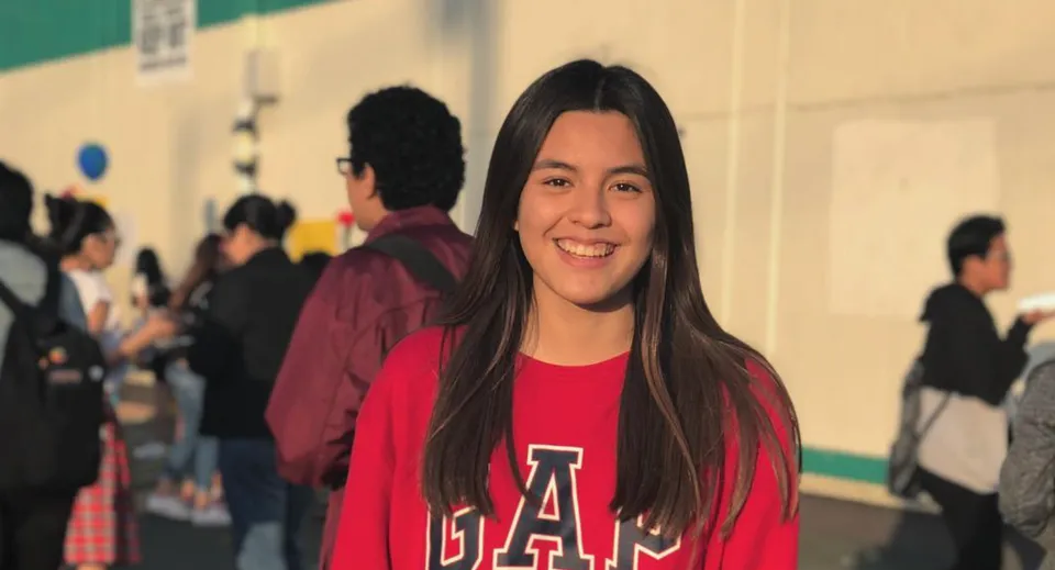 Una studentessa sorridente con una maglietta rossa GAP si trova davanti a un gruppo di persone nel campus della Downtown Magnet High School.