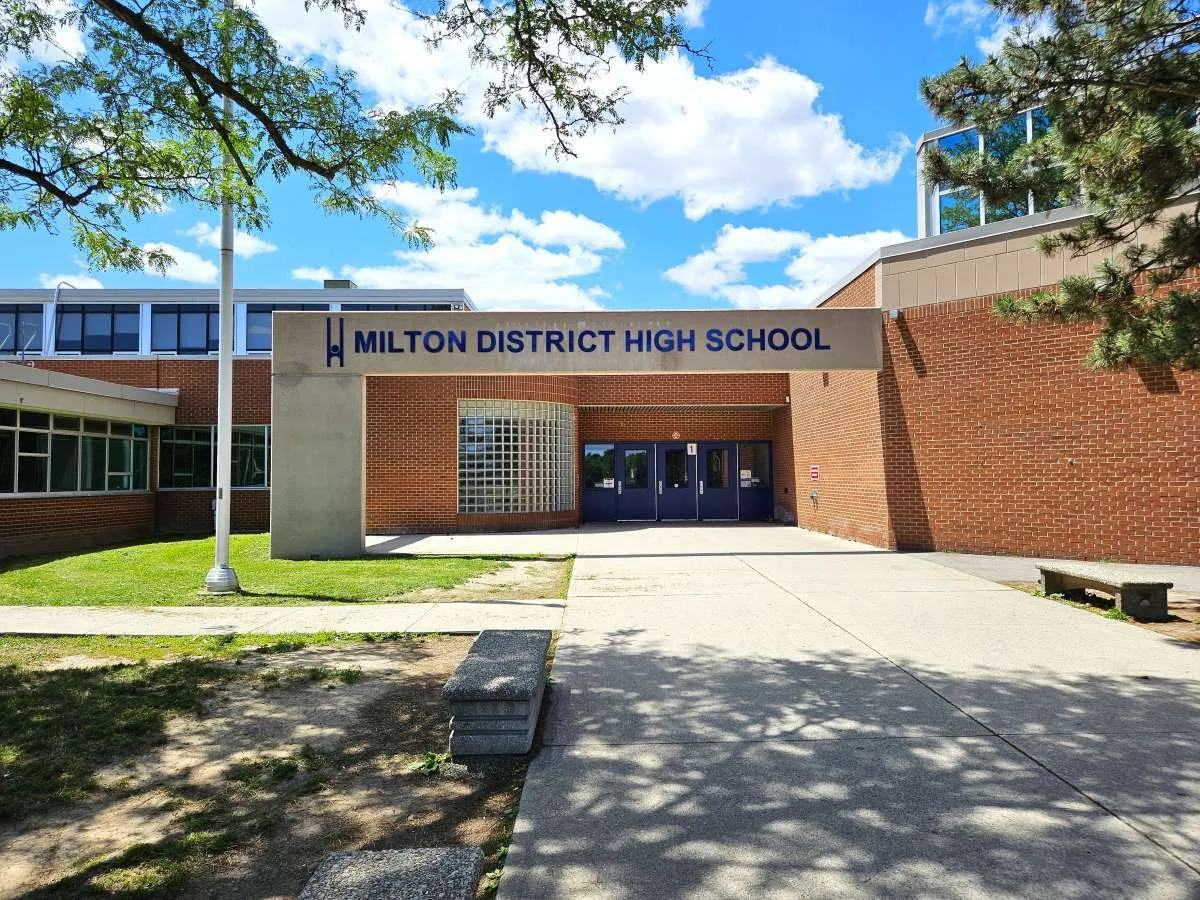 El edificio de ladrillo de la Milton District High School se encuentra rodeado de zonas de césped y árboles bajo un cielo azul con nubes.