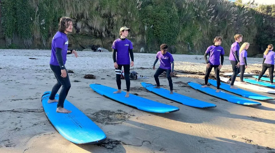 Studenti della Lake Tahoe Preparatory School in maglietta viola e muta da surf in spiaggia accanto a tavole da surf blu davanti a una foresta verde.