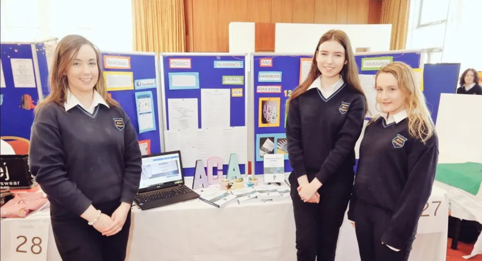 Tre studentesse in divisa scolastica sono in piedi davanti a una bacheca nell'edificio scolastico del Coláiste Mhuire Ballymote.
