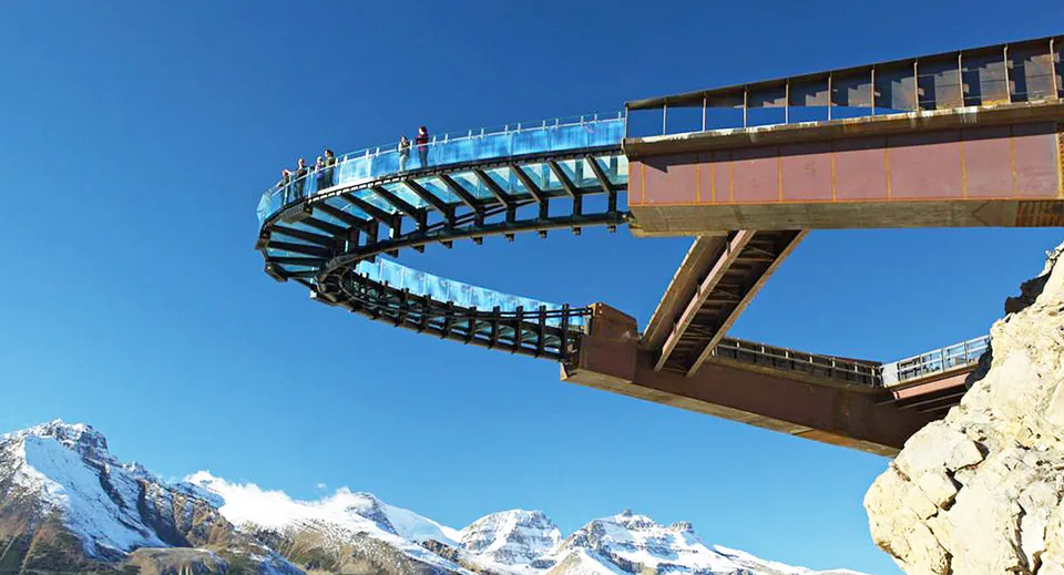 Persone su una piattaforma panoramica in vetro di fronte a cime montuose innevate.