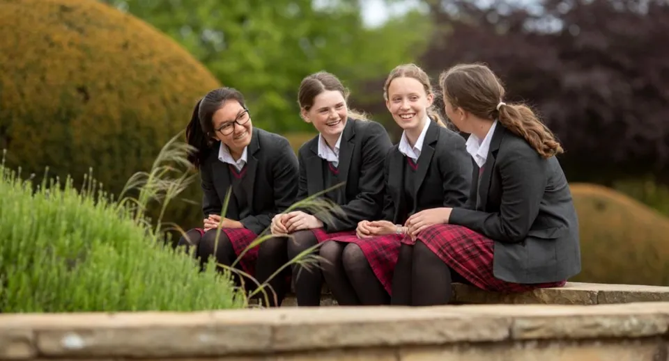 Quattro studentesse della Queen Margaret's School for Girls sono sedute su una panchina di legno nel cortile verde della scuola.