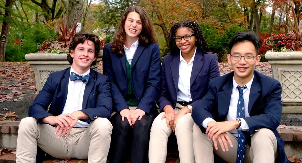 Quattro studenti siedono su una panchina di pietra all'aperto nel campus della Boston Trinity Academy davanti a uno sfondo di alberi autunnali.