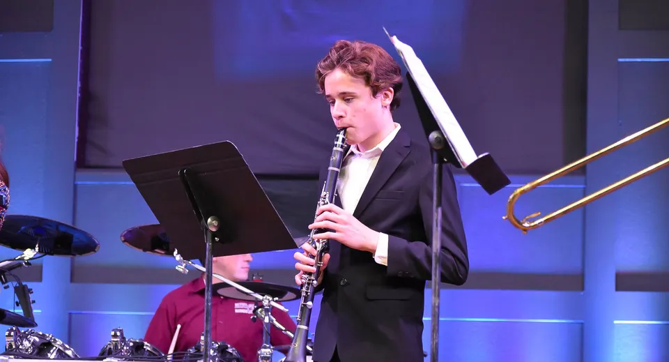 Una persona in abito elegante suona uno strumento musicale sul palco della Bethel Christian Schools con uno sfondo illuminato di blu.