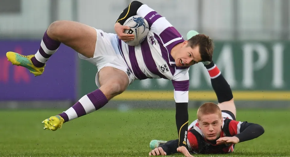 Un giocatore con una maglia a strisce viola e bianche si tuffa sopra un giocatore con la maglia rossa sul campo da gioco del Clongowes Wood College.