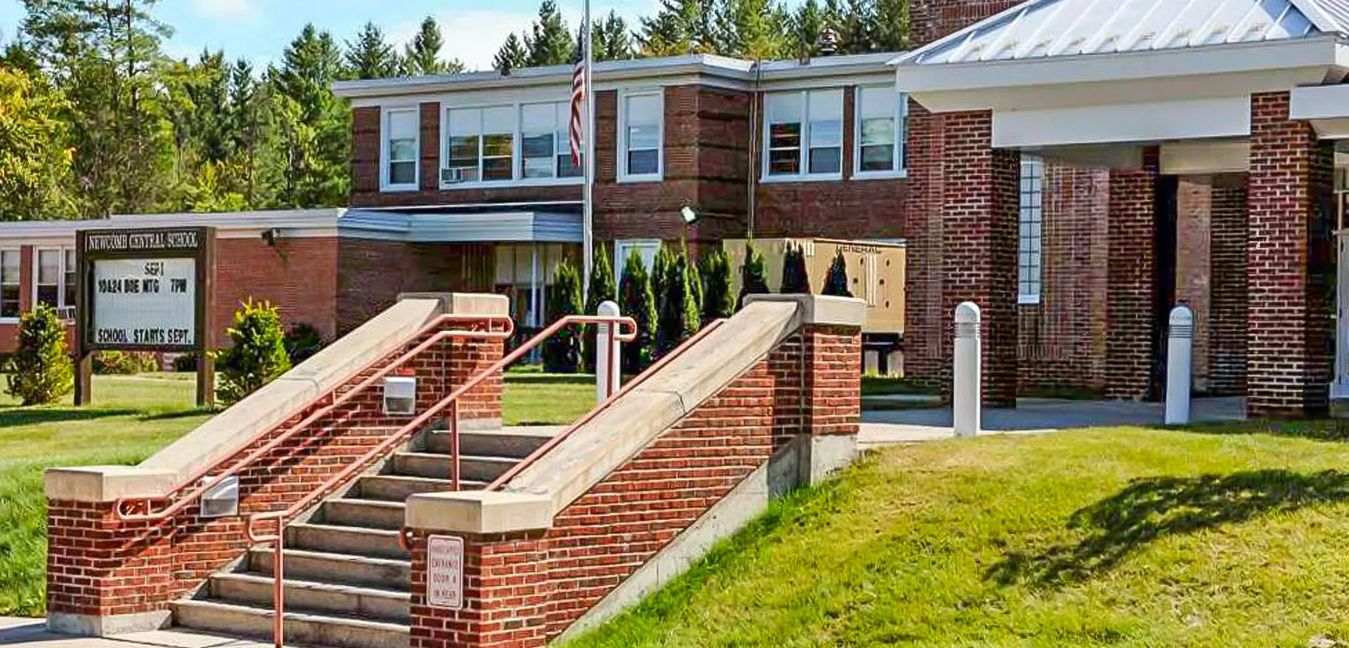 L'ingresso principale dell'edificio in mattoni nei terreni del Newcomb Central School District con scale e verde circostante.