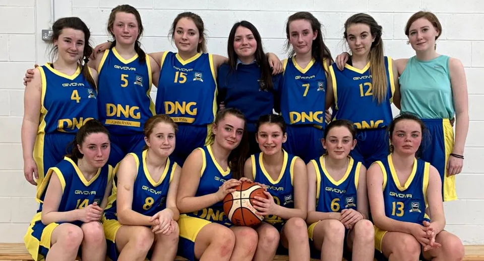 Un gruppo di giovani studentesse della Scoil Mhuire Kanturk posa come squadra di basket con un pallone davanti a una parete semplice.