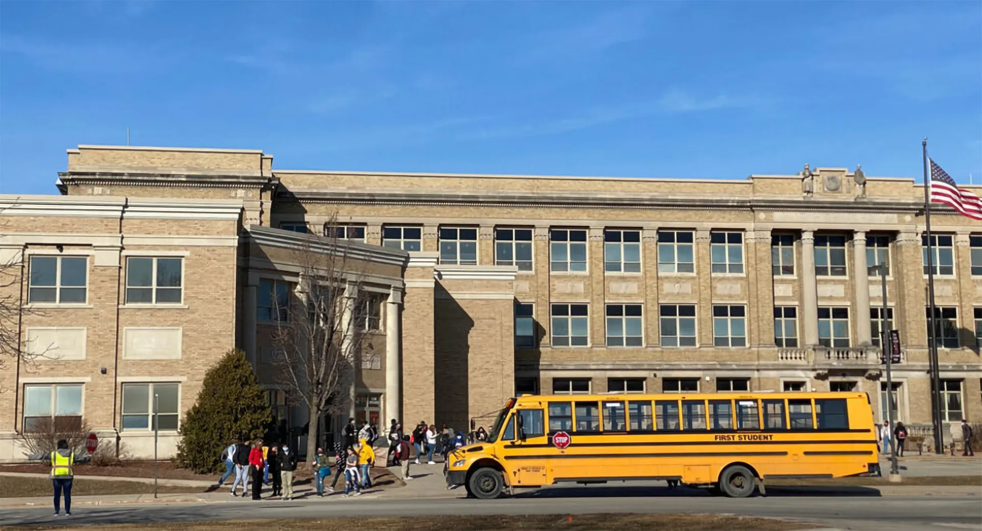 Un autobus scolastico giallo è parcheggiato davanti all'edificio del Green Bay Area Public School District con alcuni studenti all'esterno.