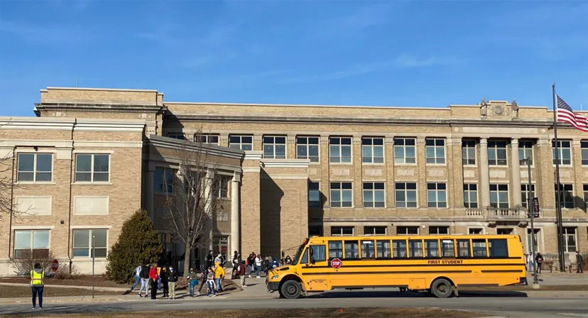 Un autobus scolastico giallo è parcheggiato davanti all'edificio del Green Bay Area Public School District con alcuni studenti all'esterno.