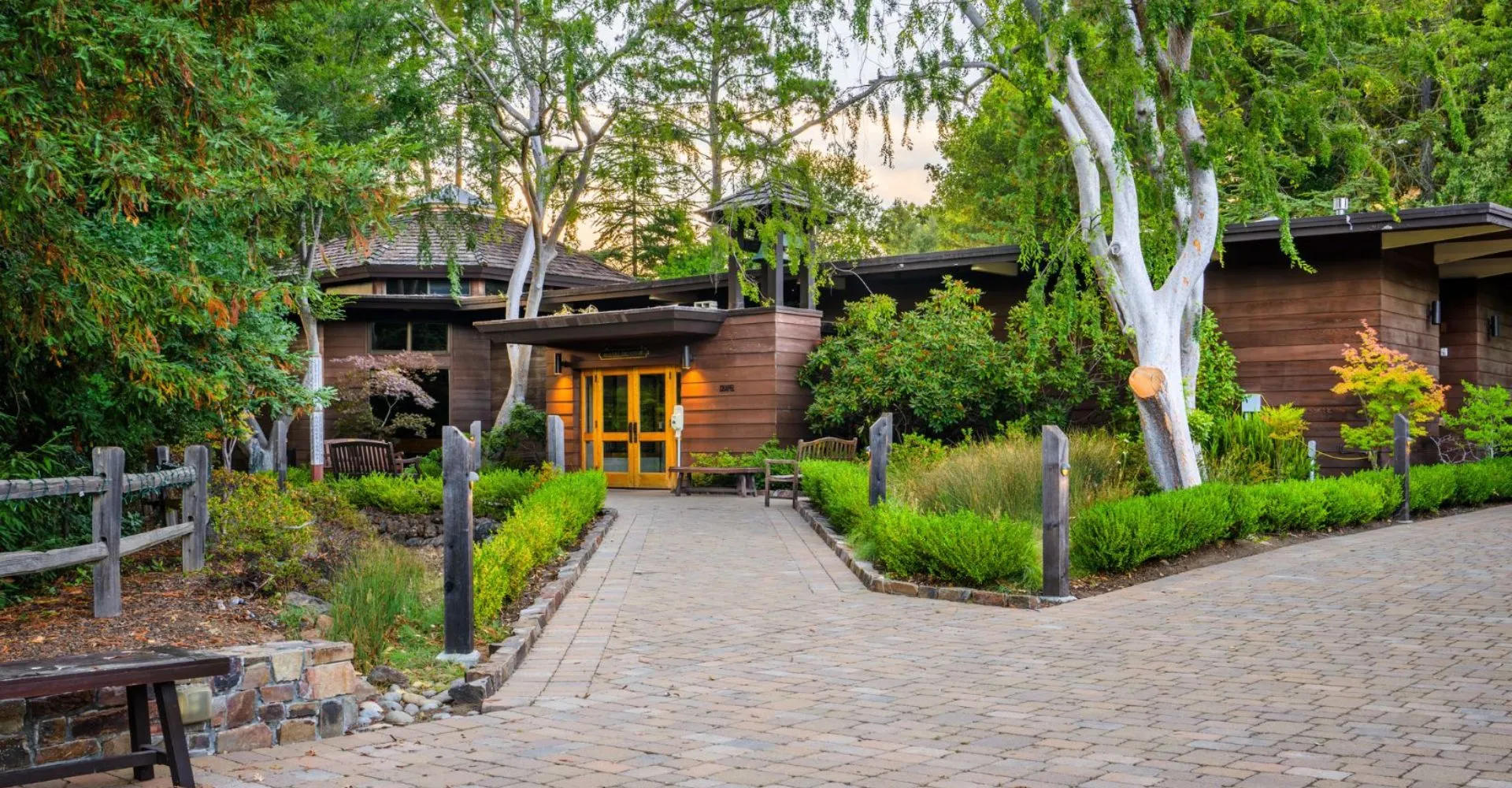 Un edificio rustico in legno con un ingresso accogliente si trova nel campus della Woodside Priory School, circondato da alberi e da un sentiero pavimentato.
