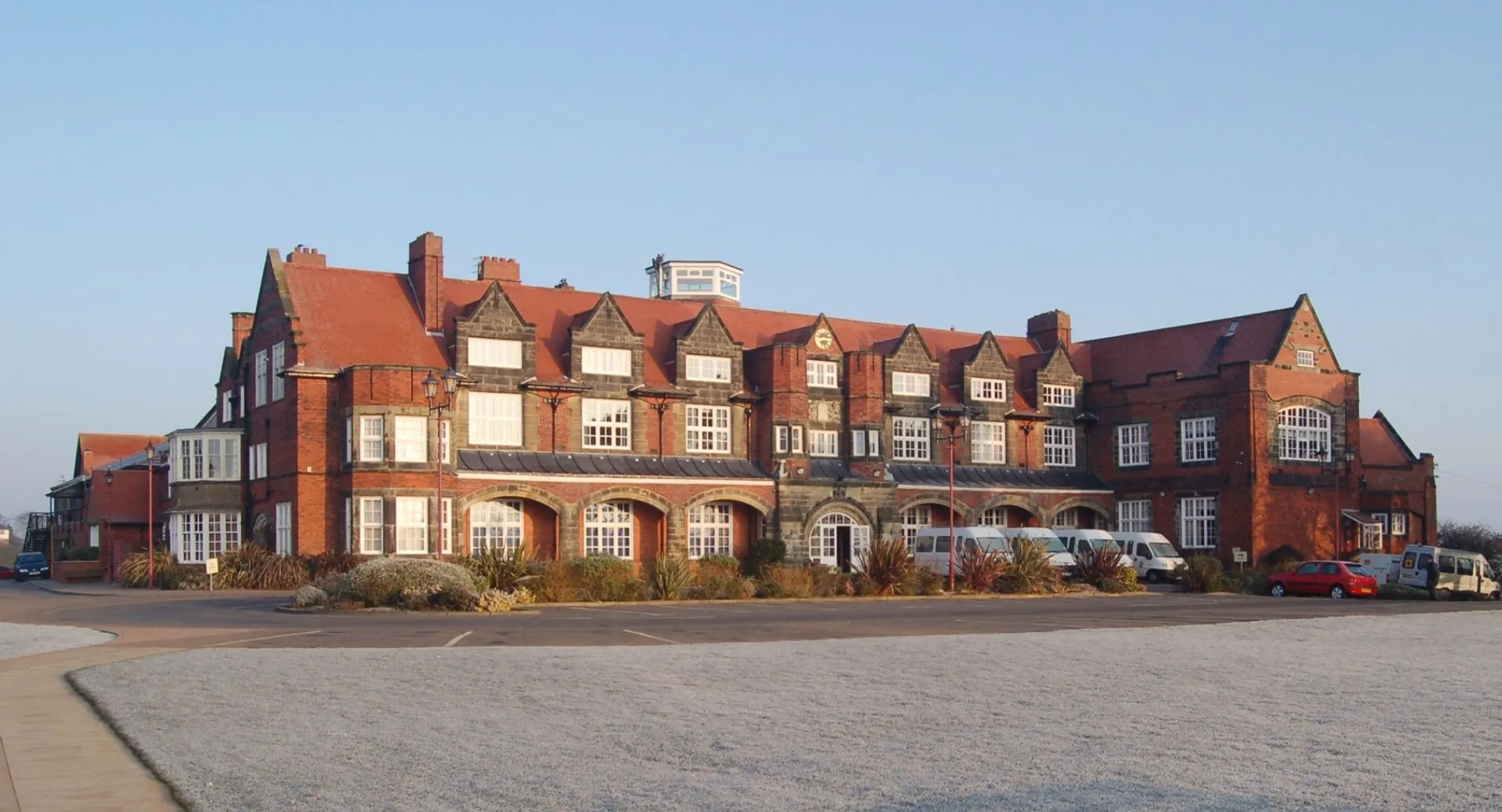 Il grande edificio in mattoni con tetto rosso e molte finestre del Scarborough College si trova davanti a un'area pavimentata con auto parcheggiate.