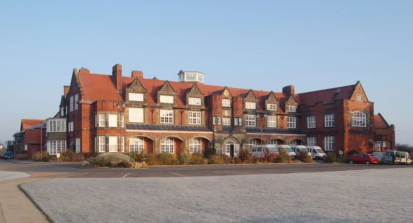 Il grande edificio in mattoni con tetto rosso e molte finestre del Scarborough College si trova davanti a un'area pavimentata con auto parcheggiate.