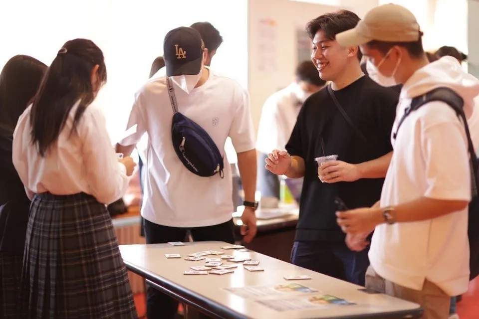 Schüler des NUCB International College stehen um einen Tisch mit kleinen Karten und unterhalten sich.