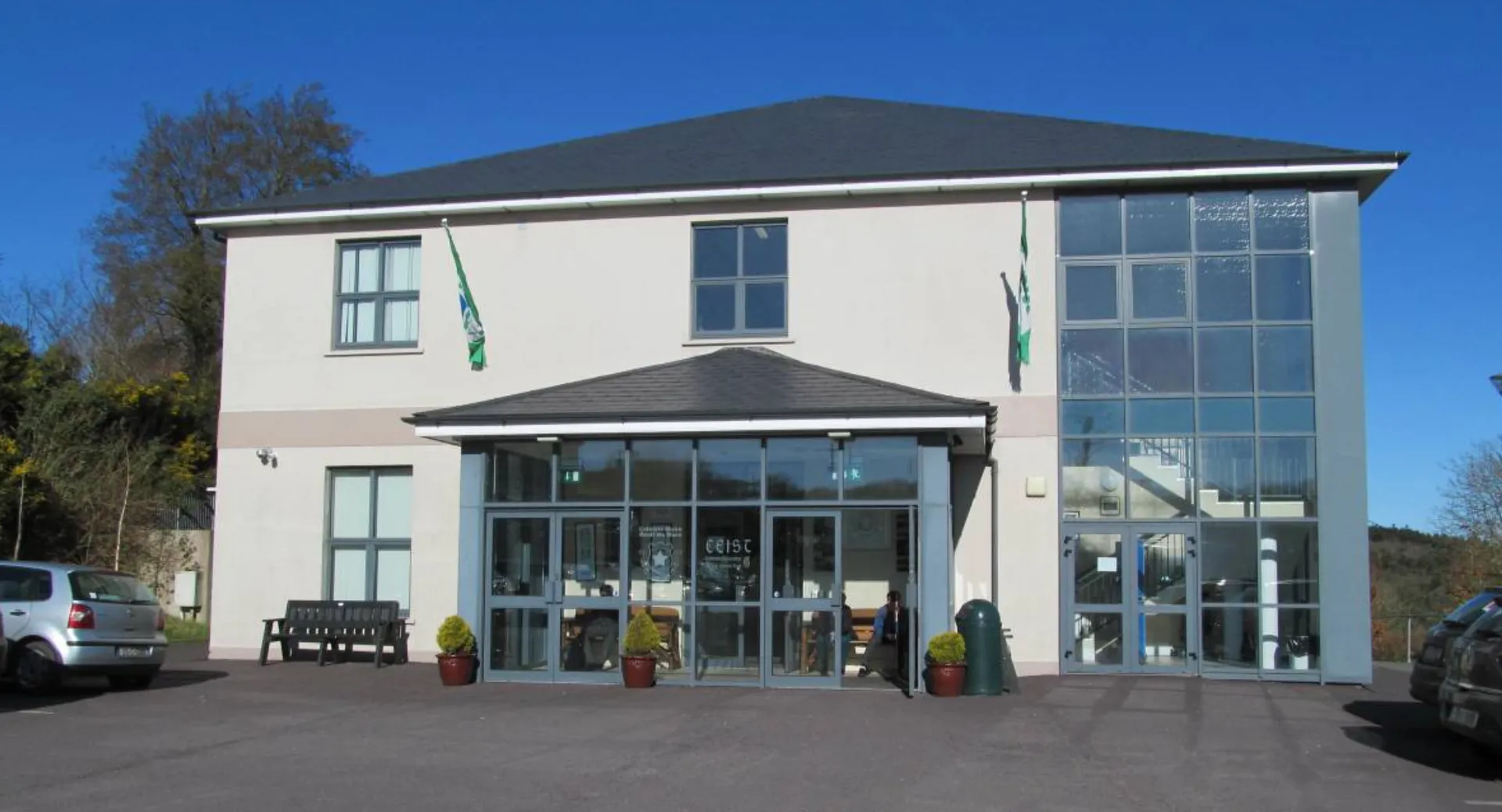 L'edificio moderno a due piani con facciata in vetro del Coláiste Muire Crosshaven è circondato da un parcheggio pavimentato e alberi.