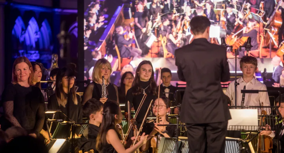Un'ampia orchestra suona sul palco sotto la direzione del direttore d'orchestra al Clifton College.