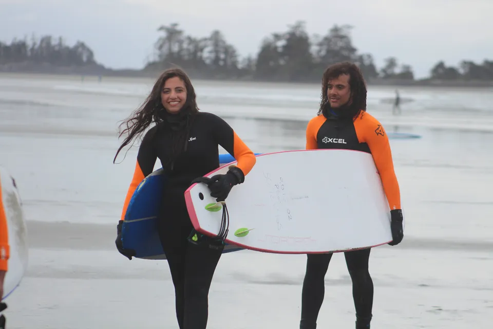 Due persone in muta con tavole da surf si trovano sulla spiaggia davanti a uno sfondo boscoso nell'area della Wellington Secondary School.