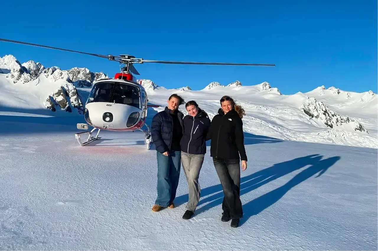 Tre studentesse davanti a un elicottero sulla neve tra montagne innevate in Nuova Zelanda