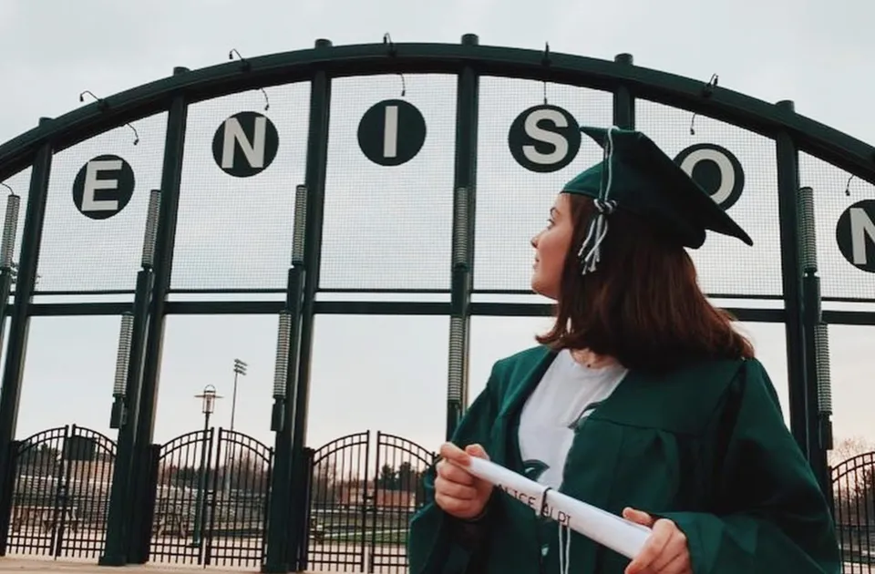 Una persona con un cappotto verde davanti al grande cancello di metallo con la scritta ENOSC nel campus della Jenison High School.