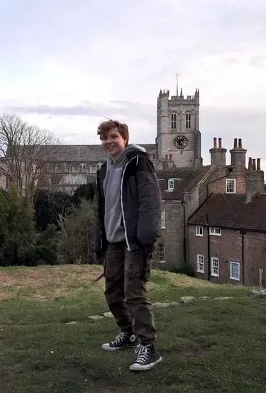Chico sonriendo en una pradera ante una iglesia medieval y casas de ladrillo