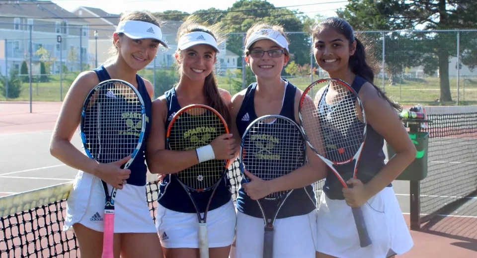 Quattro studentesse della Immaculata High School si trovano su un campo da tennis con le racchette, circondate da alberi e una recinzione.