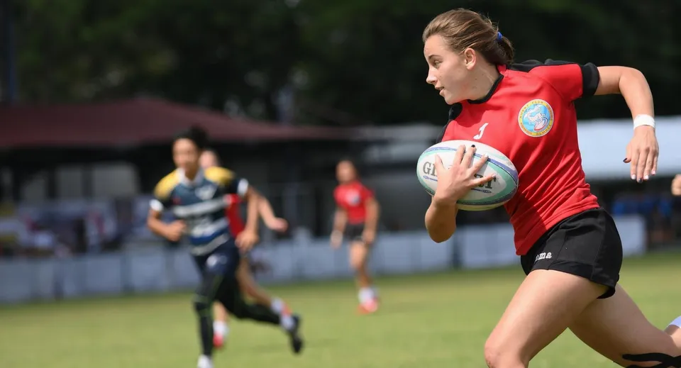 Una giocatrice di rugby in maglia rossa corre con il pallone sul campo da gioco della Mayfield School.