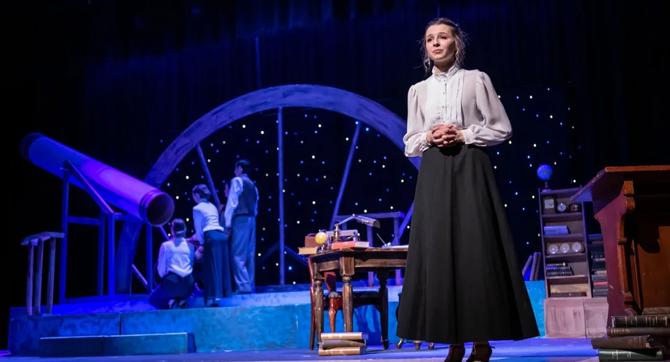 Una donna si trova sul palco della Norwell District Secondary School davanti a un arco blu con oggetti di scena sullo sfondo.