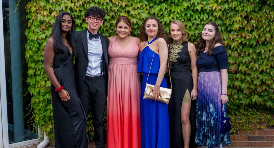 Sei persone in abito formale posano davanti a una parete coperta d'edera nel campus della Woodhouse Grove School.