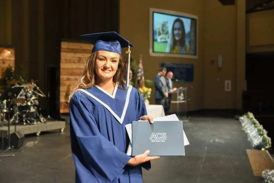 Una giovane donna in abito da diploma tiene il suo diploma nel campus della Abbotsford Christian School.