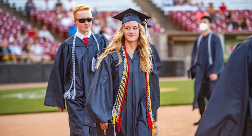 Una diplomata con toga e tocco è in piedi davanti al campus della St. Joseph High School con altri diplomati sullo sfondo.