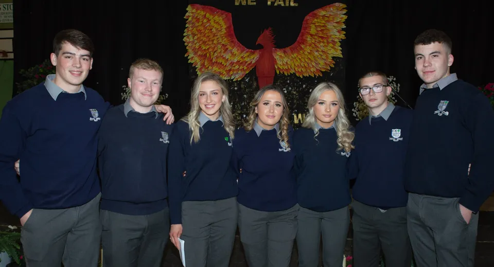 Un gruppo di studenti della Mountmellick Community School sta davanti a un grande stemma a forma di fenice su sfondo scuro.