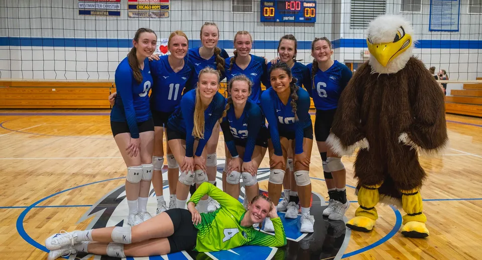 Un gruppo di pallavoliste in maglia blu posa insieme sul campo da basket della Christian Life School con una mascotte gialla.